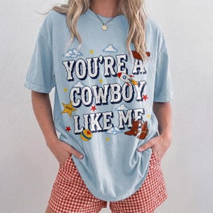 Puede incluir: Una camiseta de manga corta azul claro con el texto "YOU'RE A COWBOY LIKE ME" en blanco con un borde oscuro. El gr&aacute;fico incluye un cohete, una estrella de sheriff amarilla, una pelota colorida y botas de vaquero marrones rodeadas de estrellas y nubes.