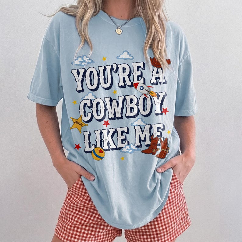 Puede incluir: Una camiseta de manga corta azul claro con el texto "YOU'RE A COWBOY LIKE ME" en blanco con un borde oscuro. El gr&aacute;fico incluye un cohete, una estrella de sheriff amarilla, una pelota colorida y botas de vaquero marrones rodeadas de estrellas y nubes.