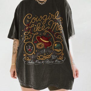 May include: A charcoal gray oversized t-shirt featuring a graphic print with the text Cowgirl Like Me, Eyes full of Stars, and Takes One to Know One. The design includes a red hat, a horseshoe, a rope border, musical notes, and floral accents.