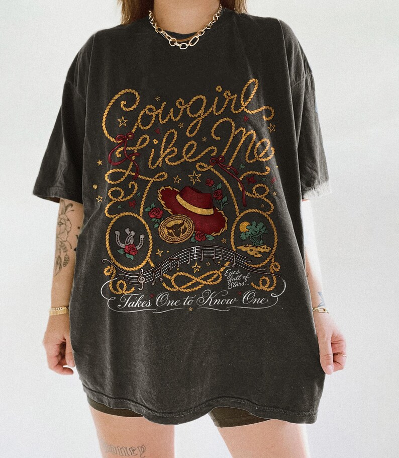 May include: A charcoal gray oversized t-shirt featuring a graphic print with the text Cowgirl Like Me, Eyes full of Stars, and Takes One to Know One. The design includes a red hat, a horseshoe, a rope border, musical notes, and floral accents.