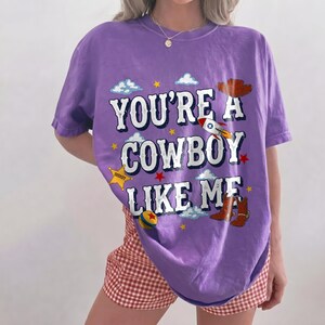 Puede incluir: Una camiseta morada de manga corta con el texto blanco "YOU'RE A COWBOY LIKE ME" e ilustraciones gr&aacute;ficas de un cohete, una insignia de sheriff, un sombrero de vaquero, botas marrones, una pelota colorida, estrellas amarillas y nubes blancas.