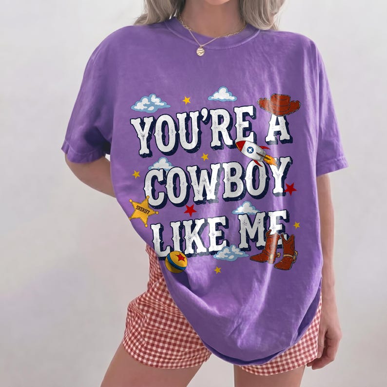 Puede incluir: Una camiseta morada de manga corta con el texto blanco "YOU'RE A COWBOY LIKE ME" e ilustraciones gr&aacute;ficas de un cohete, una insignia de sheriff, un sombrero de vaquero, botas marrones, una pelota colorida, estrellas amarillas y nubes blancas.