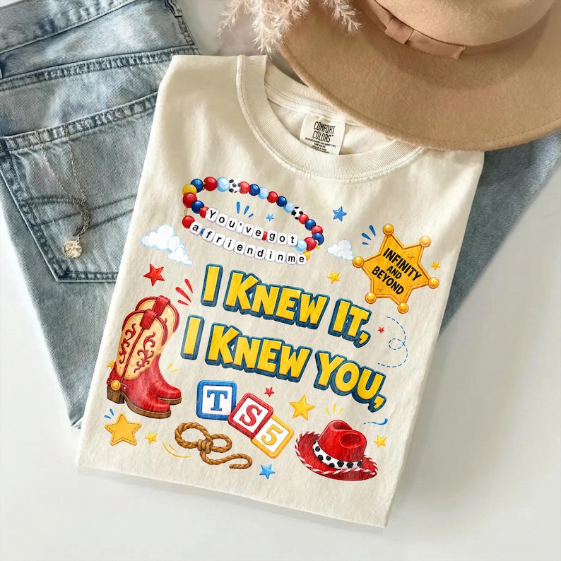 May include: A cream-colored t-shirt featuring a graphic print with a red cowboy boot, a red cowboy hat, a beaded bracelet with the text 'You've got a friend in me', a sheriff badge with 'INFINITY AND BEYOND', and the text 'I KNEW IT, I KNEW YOU, TS5'.