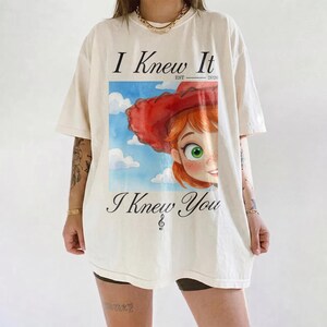K&ouml;nnte beinhalten: Ein cremefarbenes Kurzarm-T-Shirt mit einem Grafikdruck einer Figur mit roten Haaren und einem roten Hut vor einem blauen Himmel mit Wolken. Der Text lautet I Knew It EST 2026 und I Knew You, darunter befindet sich ein Violinschl&uuml;ssel.