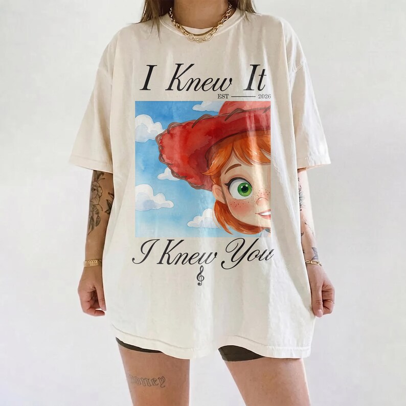 K&ouml;nnte beinhalten: Ein cremefarbenes Kurzarm-T-Shirt mit einem Grafikdruck einer Figur mit roten Haaren und einem roten Hut vor einem blauen Himmel mit Wolken. Der Text lautet I Knew It EST 2026 und I Knew You, darunter befindet sich ein Violinschl&uuml;ssel.