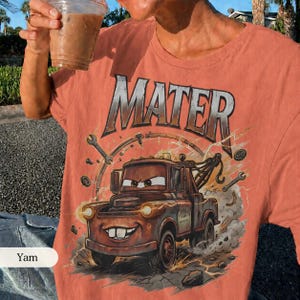 May include: Coral-colored t-shirt featuring a graphic of Mater from the movie Cars. The design includes the word "MATER" in large, silver letters above the cartoon tow truck. The shirt has a crew neck and short sleeves.