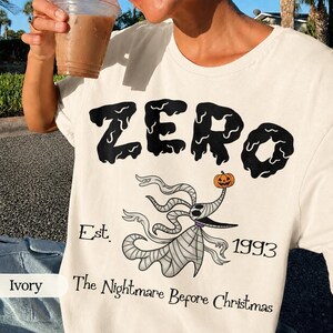 May include: Ivory-colored t-shirt featuring the word "ZERO" in a dripping black font. Below, a cartoon ghost with a pumpkin head and the text "Est. 1993 The Nightmare Before Christmas".