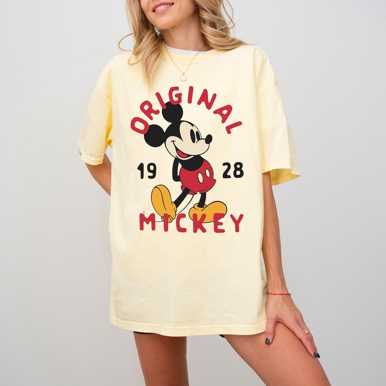 May include: A light yellow t-shirt featuring a graphic of Mickey Mouse. The design includes the word "ORIGINAL" in red, the number "1928", and the word "MICKEY" in red. Mickey Mouse is depicted in his classic outfit.