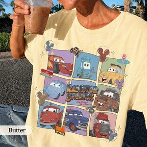 May include: A pale yellow t-shirt featuring a grid graphic of characters from the movie Cars, including Lightning McQueen, Mater, and others. The design includes the text Radiator Springs and Lightning McQueen, accented with small Mickey Mouse icons.
