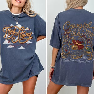 May include: A slate blue t-shirt featuring a front graphic with the text Toy Story in rope-style lettering over white clouds. The back graphic displays the text Cowgirl Like Me with a cowboy hat, rope accents, and the text Takes One to Know One.