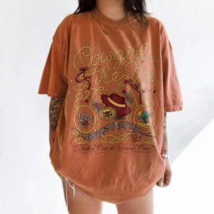 May include: An oversized burnt orange t-shirt featuring a graphic print with rope-style lettering that reads Cowgirl like Me. The design includes a cowboy hat, a horseshoe, a desert landscape, musical notes, and the text Eyes full of stars... Takes One to Know One.