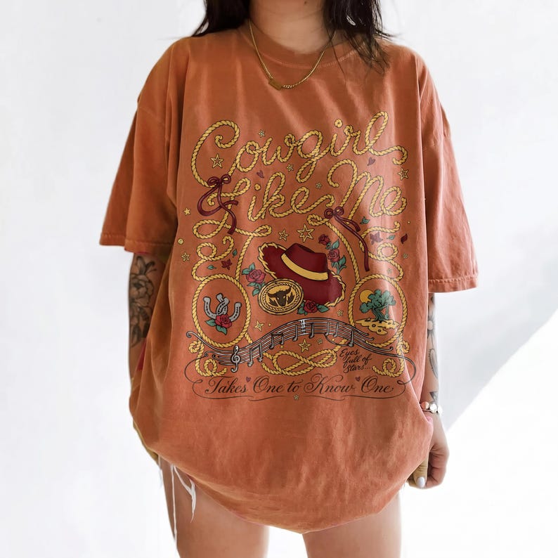 May include: An oversized burnt orange t-shirt featuring a graphic print with rope-style lettering that reads Cowgirl like Me. The design includes a cowboy hat, a horseshoe, a desert landscape, musical notes, and the text Eyes full of stars... Takes One to Know One.