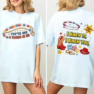 Pu&ograve; includere: Una t-shirt azzurro chiaro con una grafica sul davanti che mostra un lazo di perline e il testo "You've Got A Friend In Me". La grafica sul retro include un braccialetto di perline, una stella da sceriffo, stivali da cowboy, blocchi e un cappello.