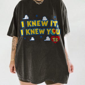May include: A charcoal gray short-sleeve t-shirt featuring the yellow text I KNEW IT, I KNEW YOU with a blue outline. The design includes small white clouds, blue stars, a colorful ball with a red star, and a red heart containing the letters TS.