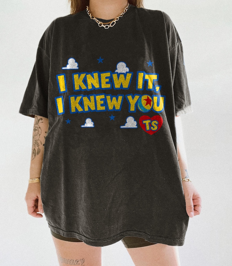 May include: A charcoal gray short-sleeve t-shirt featuring the yellow text I KNEW IT, I KNEW YOU with a blue outline. The design includes small white clouds, blue stars, a colorful ball with a red star, and a red heart containing the letters TS.
