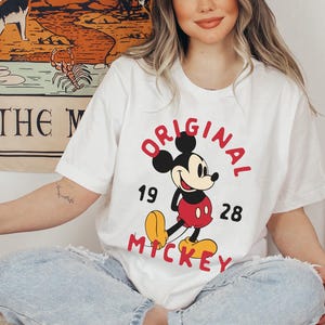 May include: White t-shirt featuring a classic Mickey Mouse design. The graphic includes the words "ORIGINAL MICKEY" in red, with the numbers "19" and "28". The shirt is a casual, vintage-inspired piece.