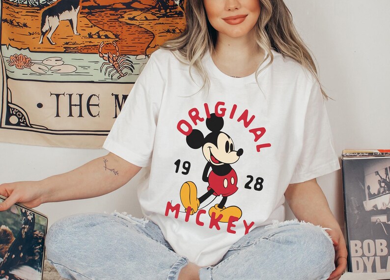 May include: White t-shirt featuring a classic Mickey Mouse design. The graphic includes the words "ORIGINAL MICKEY" in red, with the numbers "19" and "28". The shirt is a casual, vintage-inspired piece.