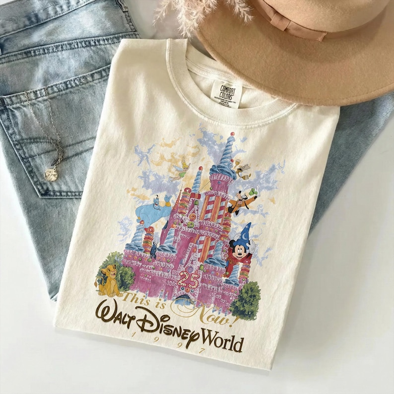May include: A cream-colored t-shirt featuring a graphic of a pink castle surrounded by various animated characters, including a blue genie, a lion, and a mouse in a wizard hat. The text reads This is Now! Walt Disney World 1971-1996.
