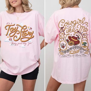 May include: A light pink t-shirt featuring a graphic on the front that reads "it's a Toy Story Baby just say yes" in rope-style lettering. The back graphic displays "Cowgirl like Me" and "Takes One to Know One" with a cowboy hat and rope motifs.