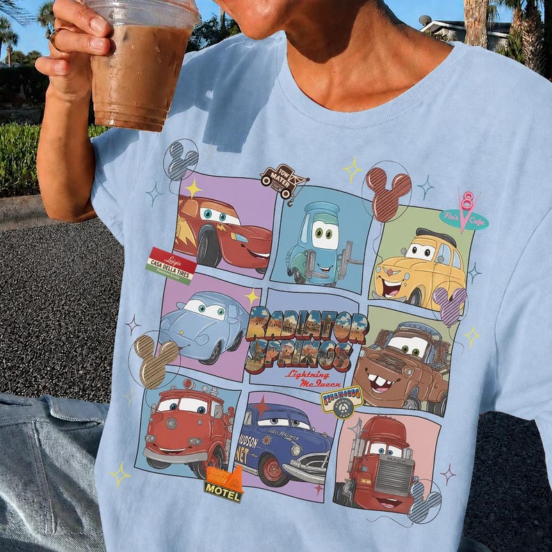 May include: A light blue t-shirt featuring a grid graphic of various animated car characters from the movie Cars. The design includes the text Radiator Springs and Lightning McQueen, surrounded by small star and balloon icons in a colorful layout.
