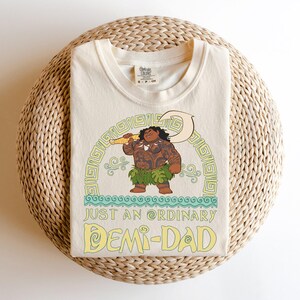 Puede incluir: Camiseta de color crema con una ilustraci&oacute;n de dibujos animados de un personaje polinesio sosteniendo un gancho. El dise&ntilde;o incluye el texto "JUST AN ORDINARY DEMI-DAD" en amarillo y verde. La camiseta est&aacute; doblada sobre una superficie circular tejida.