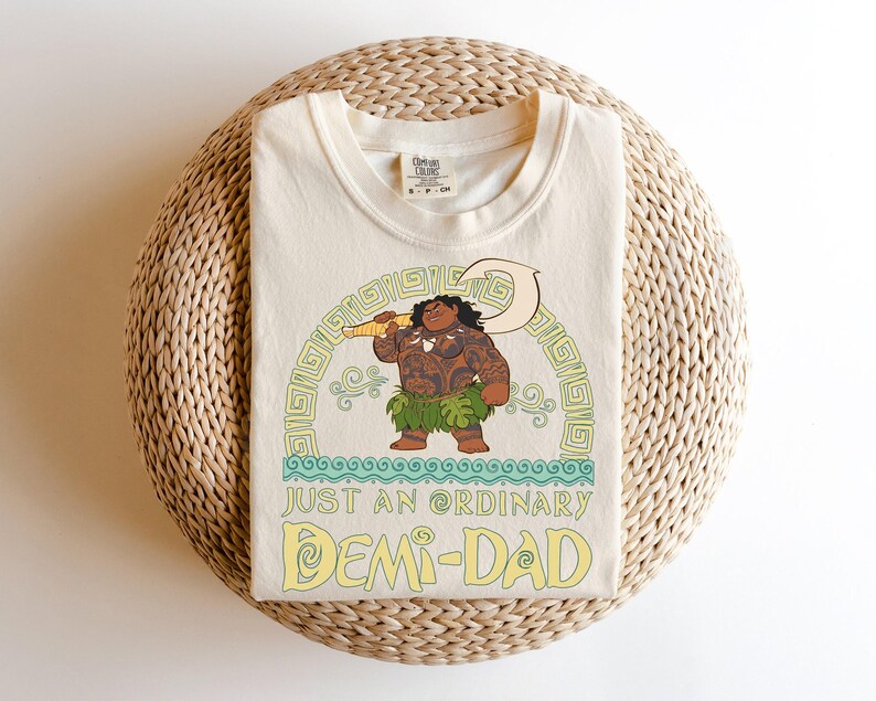 Puede incluir: Camiseta de color crema con una ilustraci&oacute;n de dibujos animados de un personaje polinesio sosteniendo un gancho. El dise&ntilde;o incluye el texto "JUST AN ORDINARY DEMI-DAD" en amarillo y verde. La camiseta est&aacute; doblada sobre una superficie circular tejida.