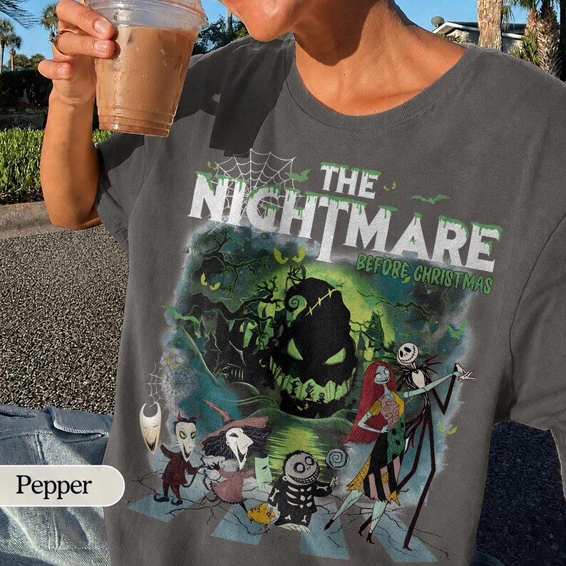 May include: Dark gray t-shirt featuring a graphic from "The Nightmare Before Christmas." The design includes characters like Jack Skellington and Sally, with the movie title in green and white. The shirt has a crew neck.