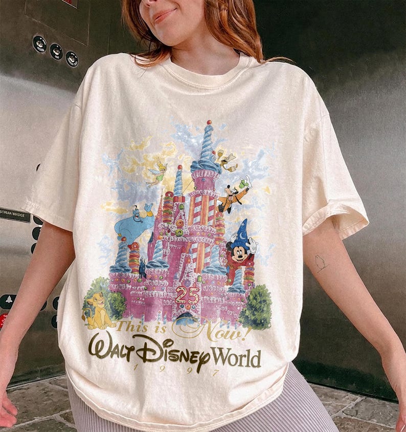May include: A cream-colored short-sleeve t-shirt featuring a colorful graphic of a castle with Disney characters including Mickey Mouse, Goofy, and Genie. The text reads 25, This is Now, and Walt Disney World 1997 in a decorative script font.