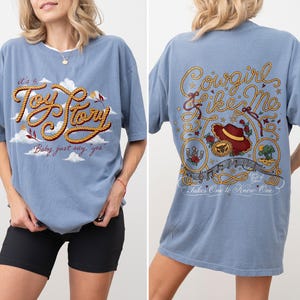 May include: A dusty blue t-shirt featuring a front graphic with the text 'it's a Toy Story' in rope-style lettering against clouds. The back graphic displays the text 'Cowgirl like Me' and 'Takes One to Know One' with a cowboy hat and rope motifs.