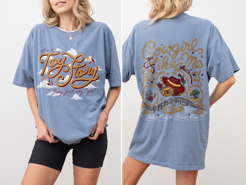 May include: A dusty blue t-shirt featuring a front graphic with the text 'it's a Toy Story' in rope-style lettering against clouds. The back graphic displays the text 'Cowgirl like Me' and 'Takes One to Know One' with a cowboy hat and rope motifs.