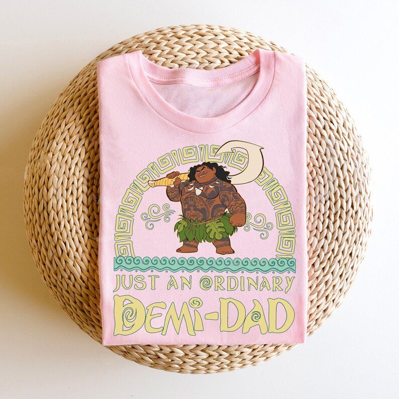 Puede incluir: Camiseta rosa claro con una ilustraci&oacute;n de dibujos animados de un personaje musculoso sosteniendo un arma. El texto "JUST AN ORDINARY DEMI-DAD" est&aacute; debajo. La camiseta est&aacute; doblada sobre una superficie tejida.