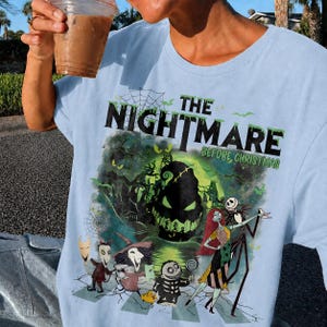 May include: Light blue t-shirt featuring a graphic from "The Nightmare Before Christmas." The design includes the film's title, Jack Skellington, Sally, and other characters in a spooky scene. The shirt is made of a soft material.