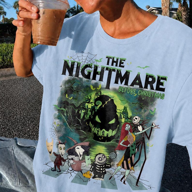 May include: Light blue t-shirt featuring a graphic from "The Nightmare Before Christmas." The design includes the film's title, Jack Skellington, Sally, and other characters in a spooky scene. The shirt is made of a soft material.