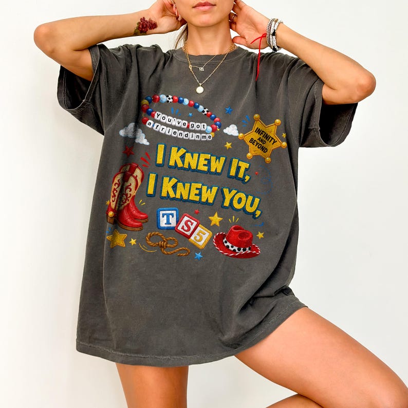 May include: A charcoal gray short-sleeve t-shirt featuring a graphic print with the text "I KNEW IT, I KNEW YOU," a beaded bracelet with "You've got a friend in me," a gold sheriff badge, red cowboy boots, a red hat, and "TS5" blocks.