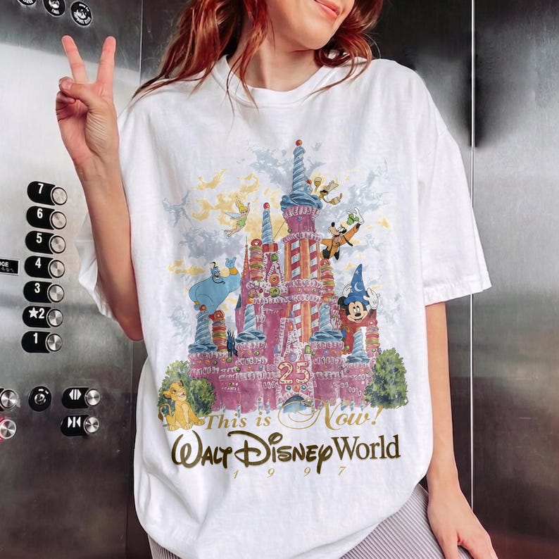 May include: A white short-sleeve t-shirt featuring a colorful graphic of a castle with Disney characters including Mickey Mouse, Genie, and Tinker Bell. The text reads 25, This is Now, Walt Disney World, and 1997, printed in a vintage style.