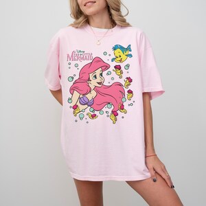 May include: A light pink t-shirt featuring a graphic of Ariel from Disney's The Little Mermaid. Ariel has long pink hair and a purple seashell top. The shirt also features Flounder and other sea creatures. The text "Disney The Little Mermaid" is above the graphic.