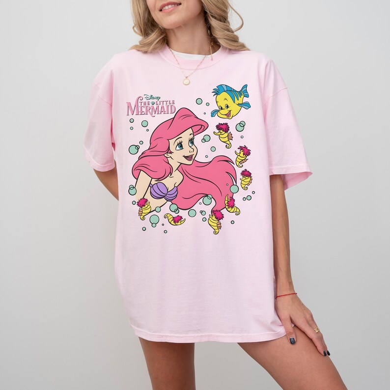 May include: A light pink t-shirt featuring a graphic of Ariel from Disney's The Little Mermaid. Ariel has long pink hair and a purple seashell top. The shirt also features Flounder and other sea creatures. The text "Disney The Little Mermaid" is above the graphic.