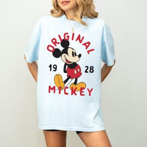 May include: Light blue t-shirt featuring a classic Mickey Mouse graphic. The design includes the word "ORIGINAL" in red, the year "1928", and the name "MICKEY" in red. The iconic cartoon character is depicted in his traditional colors.