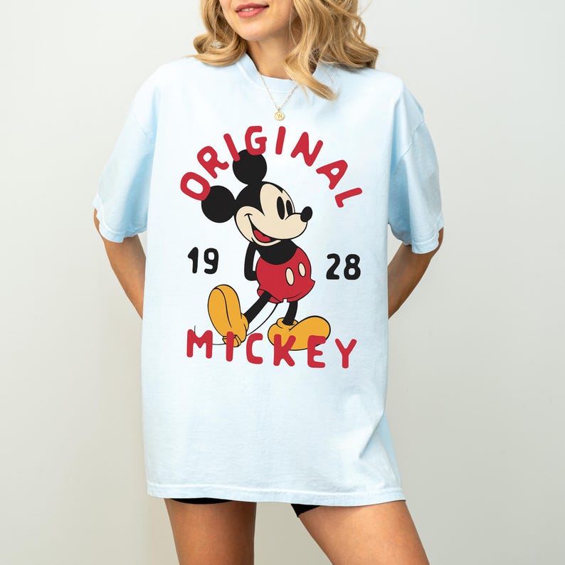 May include: Light blue t-shirt featuring a classic Mickey Mouse graphic. The design includes the word "ORIGINAL" in red, the year "1928", and the name "MICKEY" in red. The iconic cartoon character is depicted in his traditional colors.
