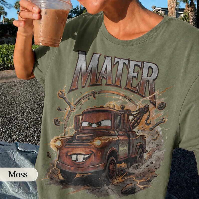 May include: Olive green t-shirt featuring a graphic of Mater from the Cars movie. The design includes the word "MATER" above the character, with tools and lightning bolts surrounding the image. The shirt is a relaxed fit.