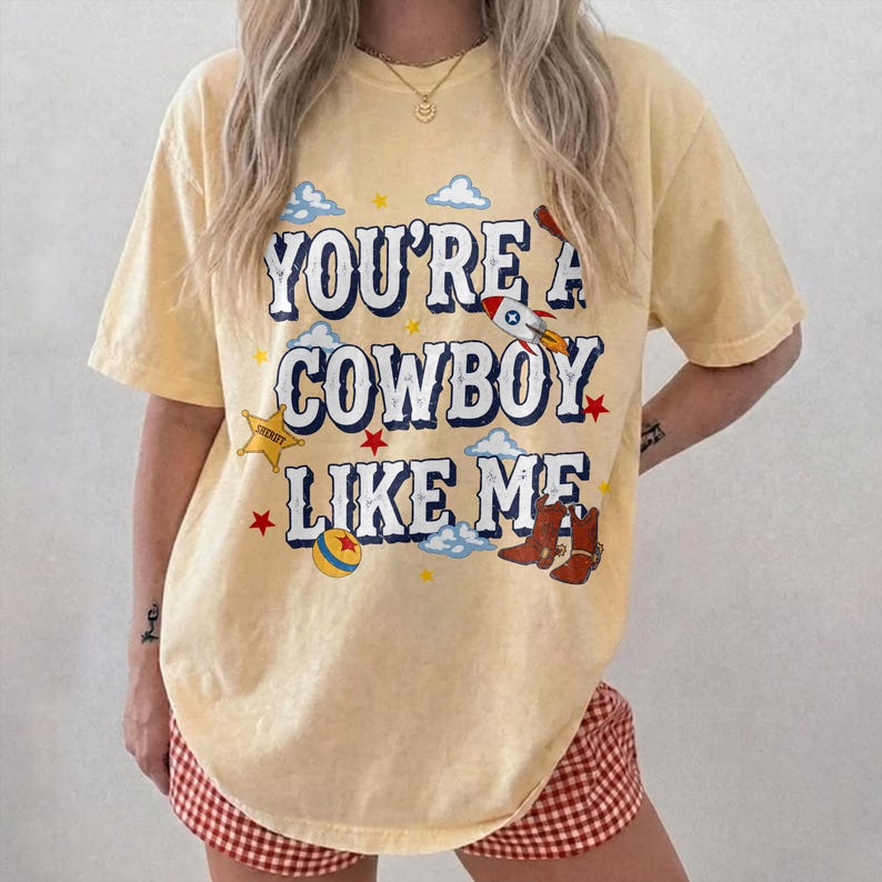 Puede incluir: Una camiseta de manga corta de color amarillo p&aacute;lido con el texto "YOU'RE A COWBOY LIKE ME" en letras azul oscuro. El gr&aacute;fico incluye ilustraciones de un cohete, una estrella de sheriff, una pelota colorida y botas vaqueras marrones.