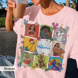 Op de afbeelding: Zachtroze T-shirt met een kleurrijke afbeelding van personages uit de Disney-film Moana. Het ontwerp bevat Moana, Maui, Pua, Hei Hei en anderen. Het shirt heeft een ronde hals en korte mouwen.