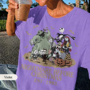 May include: Violet-colored t-shirt featuring a graphic from "The Nightmare Before Christmas." The design includes characters like Jack Skellington, Sally, and Oogie Boogie, along with pumpkins and the text "NIGHTMARE BEFORE CHRISTMAS EST. 1993."