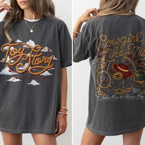 May include: A charcoal gray short-sleeve t-shirt featuring a front graphic with the text Toy Story in rope-style lettering over white clouds. The back graphic displays the text Cowgirl Like Me and Takes One To Know One with a cowboy hat.