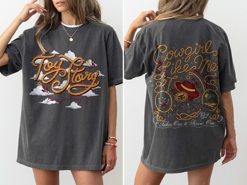 May include: A charcoal gray short-sleeve t-shirt featuring a front graphic with the text Toy Story in rope-style lettering over white clouds. The back graphic displays the text Cowgirl Like Me and Takes One To Know One with a cowboy hat.