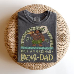 Puede incluir: Camiseta gris oscuro con una ilustraci&oacute;n de dibujos animados de un personaje sosteniendo un arma. El texto "JUST AN ORDINARY DEMI-DAD" est&aacute; en dorado y turquesa. La camiseta est&aacute; doblada sobre una superficie circular tejida.