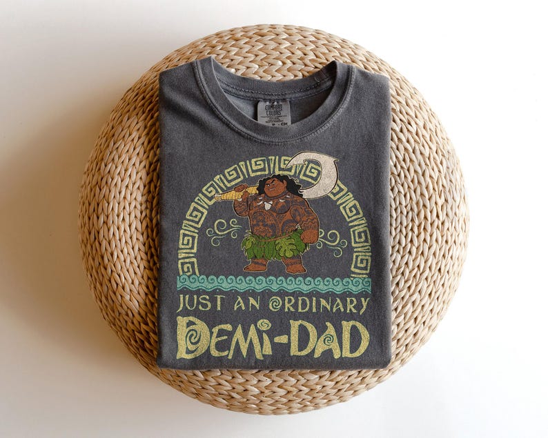 Puede incluir: Camiseta gris oscuro con una ilustraci&oacute;n de dibujos animados de un personaje sosteniendo un arma. El texto "JUST AN ORDINARY DEMI-DAD" est&aacute; en dorado y turquesa. La camiseta est&aacute; doblada sobre una superficie circular tejida.