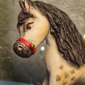May include: A detailed, hand-painted ceramic horse figurine. The horse has a cream-colored body with brown spots, a dark brown mane, and a red bridle with a gold bell. The figurine is a decorative item.