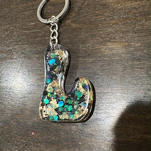 May include: A clear resin keychain in the shape of the letter "L". The resin is filled with gold flakes, teal and black glitter. The keychain has a silver-colored metal ring and chain.