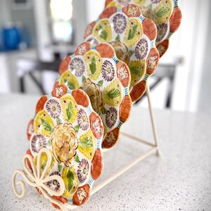 May include: A set of decorative plates with a floral design, displayed on a cream-colored metal stand. The plates feature a scalloped edge with orange, yellow, and purple floral patterns. The stand has a decorative, scrolled base.